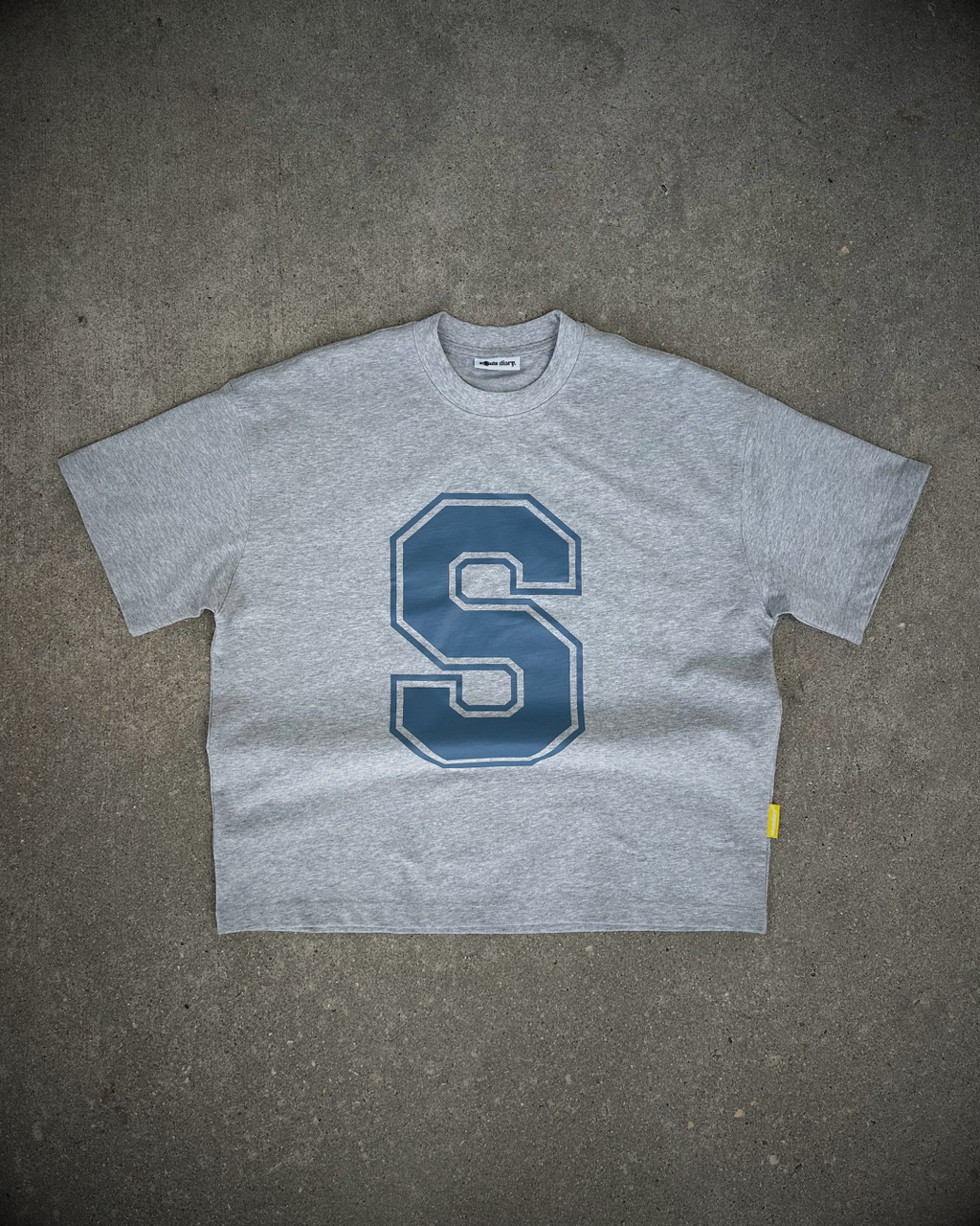 A heather gray short-sleeve t-shirt with a large blue varsity-style letter 'S' printed on the chest, laid flat on a textured gray concrete surface.
