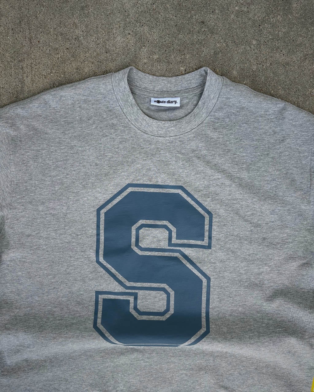 A heather gray crewneck sweatshirt featuring a large navy blue block letter 'S' outlined in white on the front, laid flat on a textured gray surface.