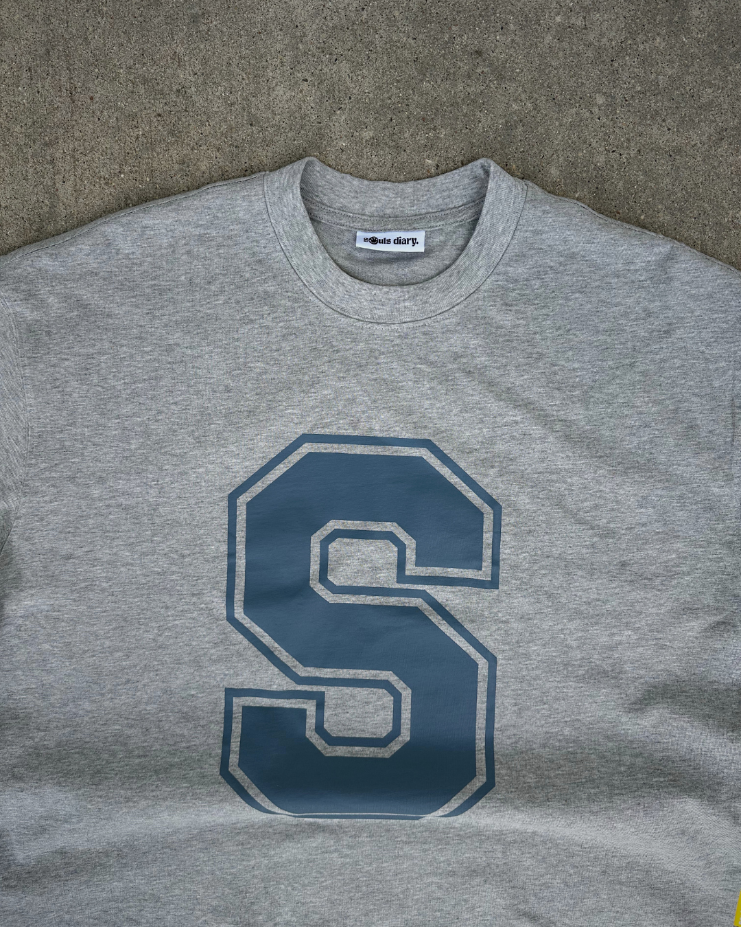 A heather gray crewneck sweatshirt featuring a large navy blue block letter 'S' outlined in white on the front, laid flat on a textured gray surface.