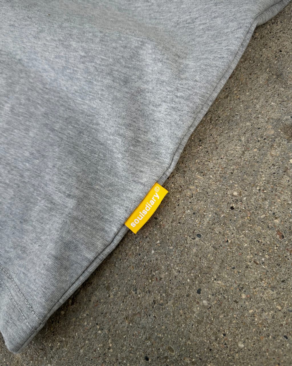 Close-up of a heather gray fabric item laid on a concrete surface, with a small yellow fabric tag reading 'soulsdiary' attached near the seam.
