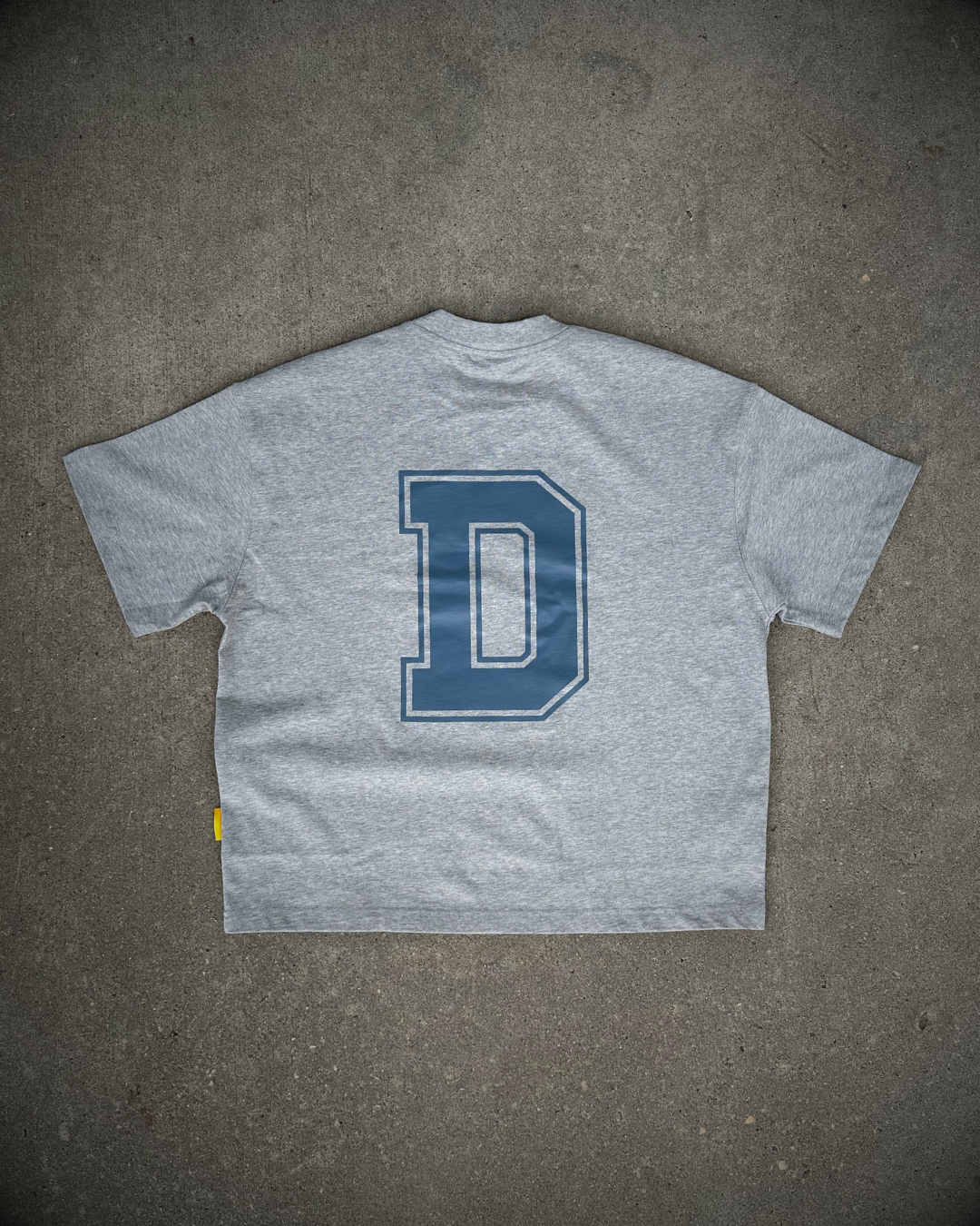 A heather gray short-sleeve t-shirt laid flat on a textured gray concrete surface, featuring a large blue block letter 'D' outlined in white on the back.